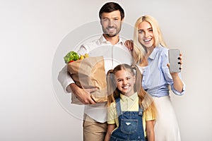 Family Showing Empty Cellphone Screen Holding Grocery Bag, Studio Shot