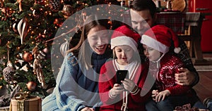 Family sit near decorated Christmas Tree having fun using smartphone