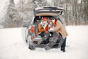 Family sitting in open car's trunk at winter park and drink a tea