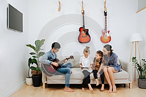 Family sitting on a sofa. Mom and kids looking on a tablet, dad plays guitar