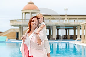 Family. A smiling mother and daughter hugging each other. Outdoor. In the background is a complex with a swimming pool. Concept of