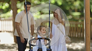A family spending time in the park - smiling happy parents swinging their daughter on a swing