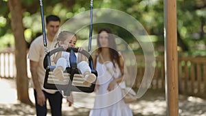 A family spending time in the park - smiling happy parents swinging their daughter on a swing in slow motion