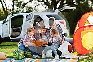 Family spending time together. Mother reading book outdoor with kids against their suv car