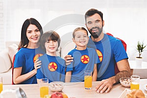 A family of superheroes sit at a table. The incredibles are posing in a bright room