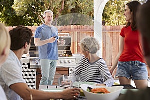 Family at a table outdoors turn to dad standing by barbecue