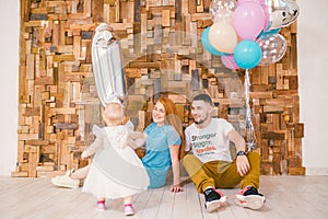 Family of three celebrates daughter`s birthday one year inside the room sitting on the floor against the background of the wooden