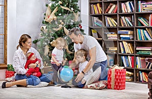 Family with three children beside Christmas tree at home. Kids and father examining globe, mom is holding baby girl