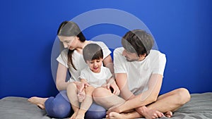 Family of three dad mom son sitting on the bed against a blue wall