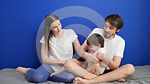 Family of three dad mom son sitting on the bed against a blue wall