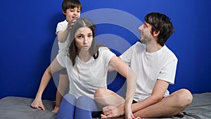 Family of three dad mom son sitting on the bed against a blue wall