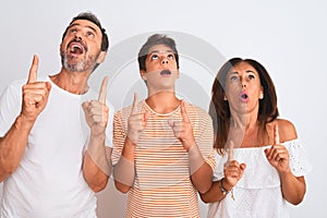 Family of three, mother, father and son standing over white isolated background amazed and surprised looking up and pointing with