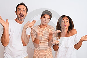 Family of three, mother, father and son standing over white isolated background clueless and confused expression with arms and