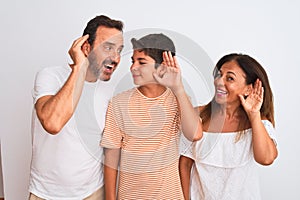 Family of three, mother, father and son standing over white isolated background smiling with hand over ear listening an hearing to