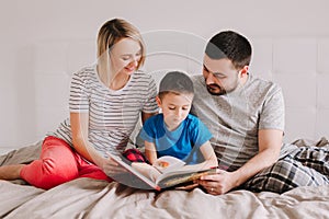 Family of three people sitting on bed in bedroom reading book. Mother, father and boy son at home spending time together. Parents
