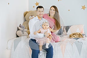 family of three sitting on a baby bed in the nursery and hugging each other and making big toothy smiles, looking at the camera