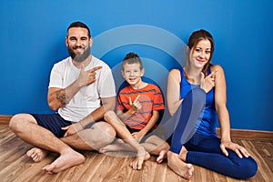 Family of three sitting on the floor at home cheerful with a smile of face pointing with hand and finger up to the side with happy
