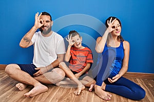 Family of three sitting on the floor at home doing ok gesture with hand smiling, eye looking through fingers with happy face