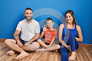 Family of three sitting on the floor at home showing and pointing up with fingers number five while smiling confident and happy