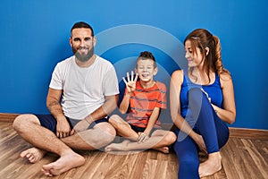 Family of three sitting on the floor at home showing and pointing up with fingers number four while smiling confident and happy