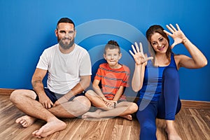Family of three sitting on the floor at home showing and pointing up with fingers number ten while smiling confident and happy