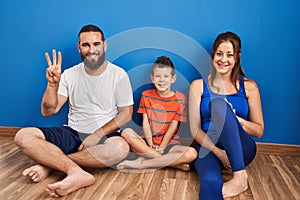 Family of three sitting on the floor at home showing and pointing up with fingers number three while smiling confident and happy