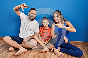 Family of three sitting on the floor at home smiling making frame with hands and fingers with happy face