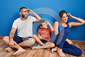 Family of three sitting on the floor at home very happy and smiling looking far away with hand over head