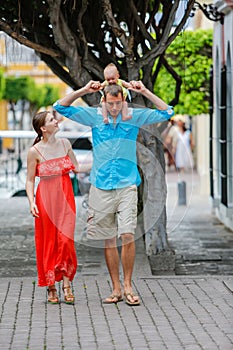 A family of three are walking. Baby girl is sitting on her fathers shoulders.