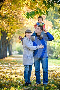 Family of three walks in the autumn park holding hands. happy father carrying son with maple leaves. Mother embrace her cute boy