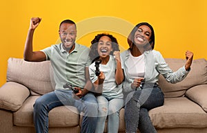 Family time activities. Happy black parents and daughter playing video games, sitting on sofa over yellow background