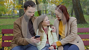 Family together happy man father woman mother child kid girl little daughter outdoors park on bench dad mom baby using