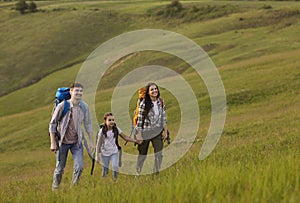A family of tourists hiking ckpacks is walking along the grass on a hill in the mountains.