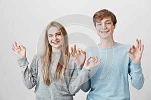 Family unity looks like this. Charming friendly siblings smiling broadly, standing near and showing ok gesture with