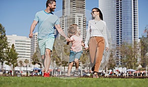 Family in urban skyline on park lawn. Happy parents holding hands with son and walking in sunny summer citi street