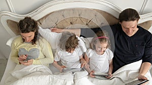 Family is Using Mobile Devices Lying in the Bed During Weekend at Home. Two Kids is Holding Tablets. Pretty Wife with