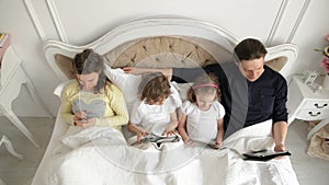 Family is Using Mobile Devices Together in the Bedroom During Weekend at Home. Two Kids is Holding Tablets. Pretty Wife