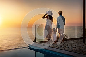 A family on vacation stands by the pool and enjoys the sunset over the sea