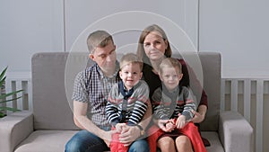 Family video portrait of mom, dad and two twins brothers toddlers. They are siting on the sofa and looking at camera.