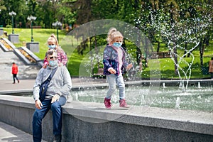 Family walk in the park in medical masks. A cute little blonde baby in a medical mask runs along the side of the fountain and