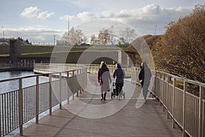 Family is walking in park. Three women walk across bridge over water. People in city in fall. Details of park architecture