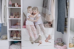 Family wardrobe. Happy twin sisters on shelve in closet with natural organic clothes in cozy dressing room play joyfully