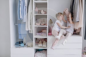 Family wardrobe. Happy twin sisters on shelve in closet with natural organic clothes in cozy dressing room play joyfully