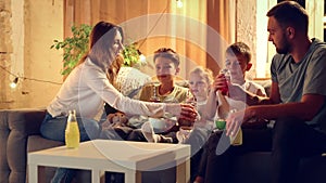 Family watching tv at home. Young woman, man and children sitting on sofa in living room, eating snacks and watching