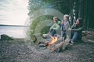 Family weekend outdoor. Father, mother and child sitting near bonfire in forest near lake. Local travel, camping, hiking lifestyle