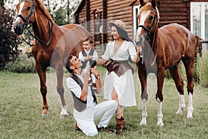 A family in white clothes with their son stand near two beautiful horses in nature. A stylish couple with a child are