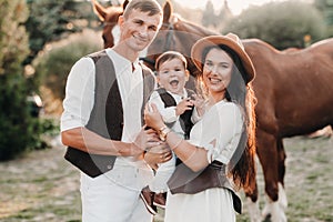 A family in white clothes with their son stand near two beautiful horses in nature. A stylish couple with a child are photographed