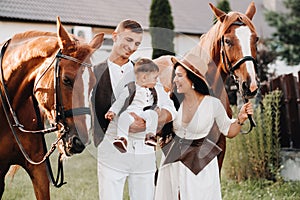 A family in white clothes with their son stand near two beautiful horses in nature. A stylish couple with a child are photographed