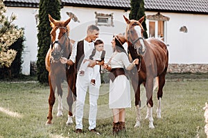 A family in white clothes with their son stand near two beautiful horses in nature. A stylish couple with a child are photographed