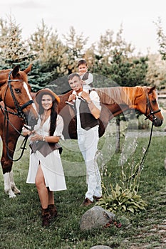 A family in white clothes with their son stand near two beautiful horses in nature. A stylish couple with a child are photographed